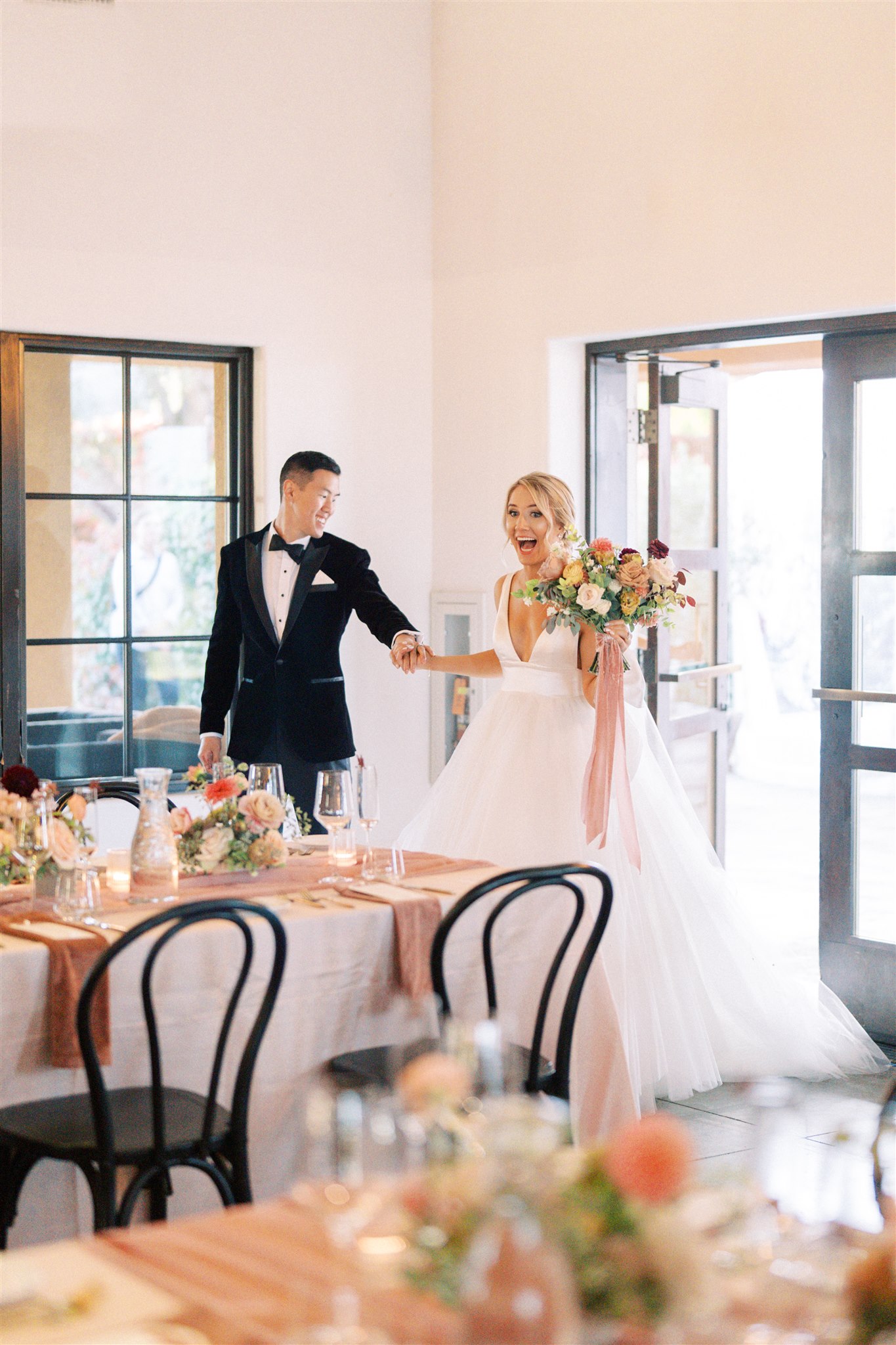 A bride in a white dress holding a bouquet walks into a decorated reception room, smiling and holding hands with a man in a tuxedo.