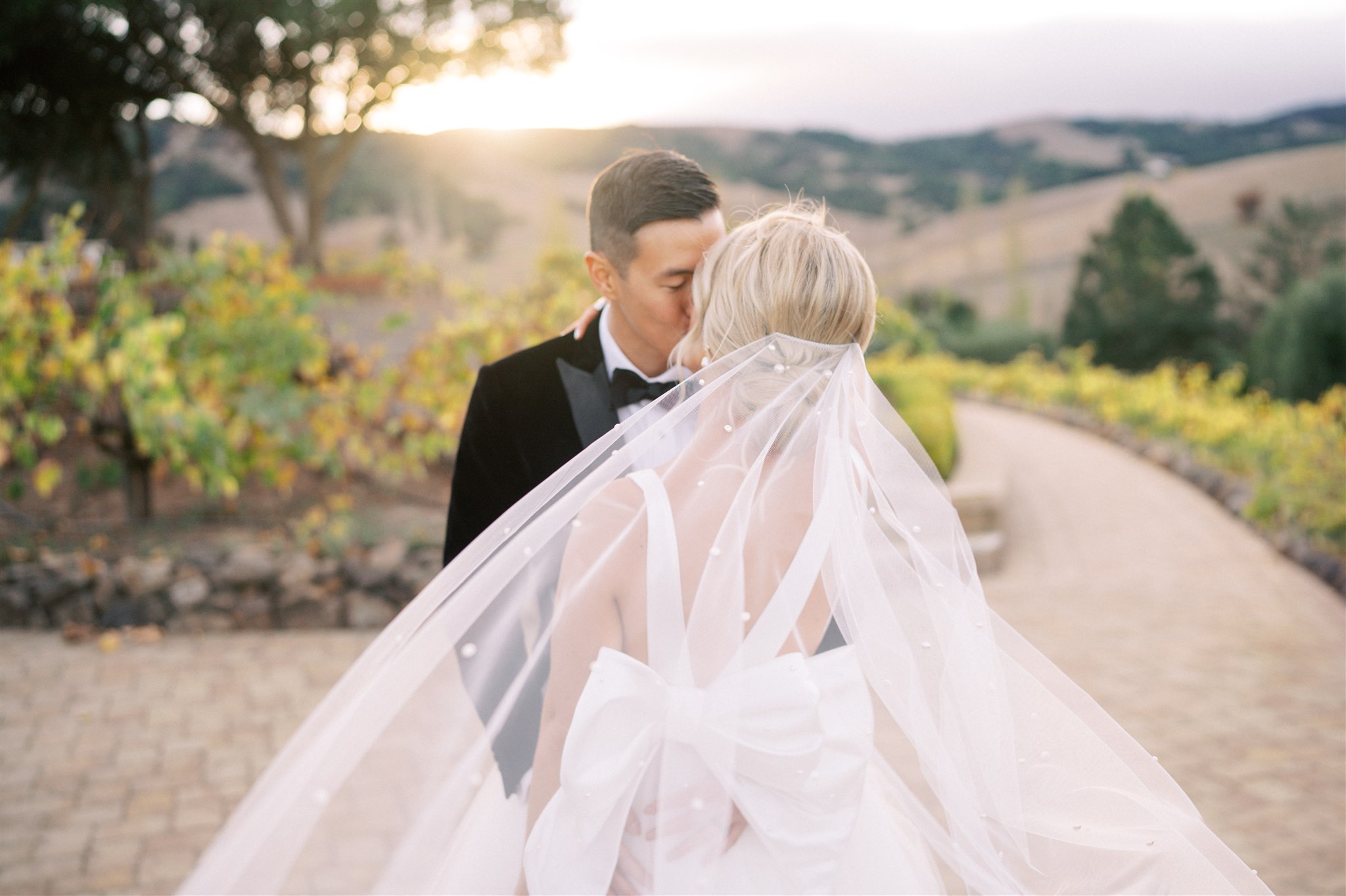 A bride and groom kiss outdoors at sunset, with vineyards and hills in the background. The bride’s veil and dress are visible in the foreground.