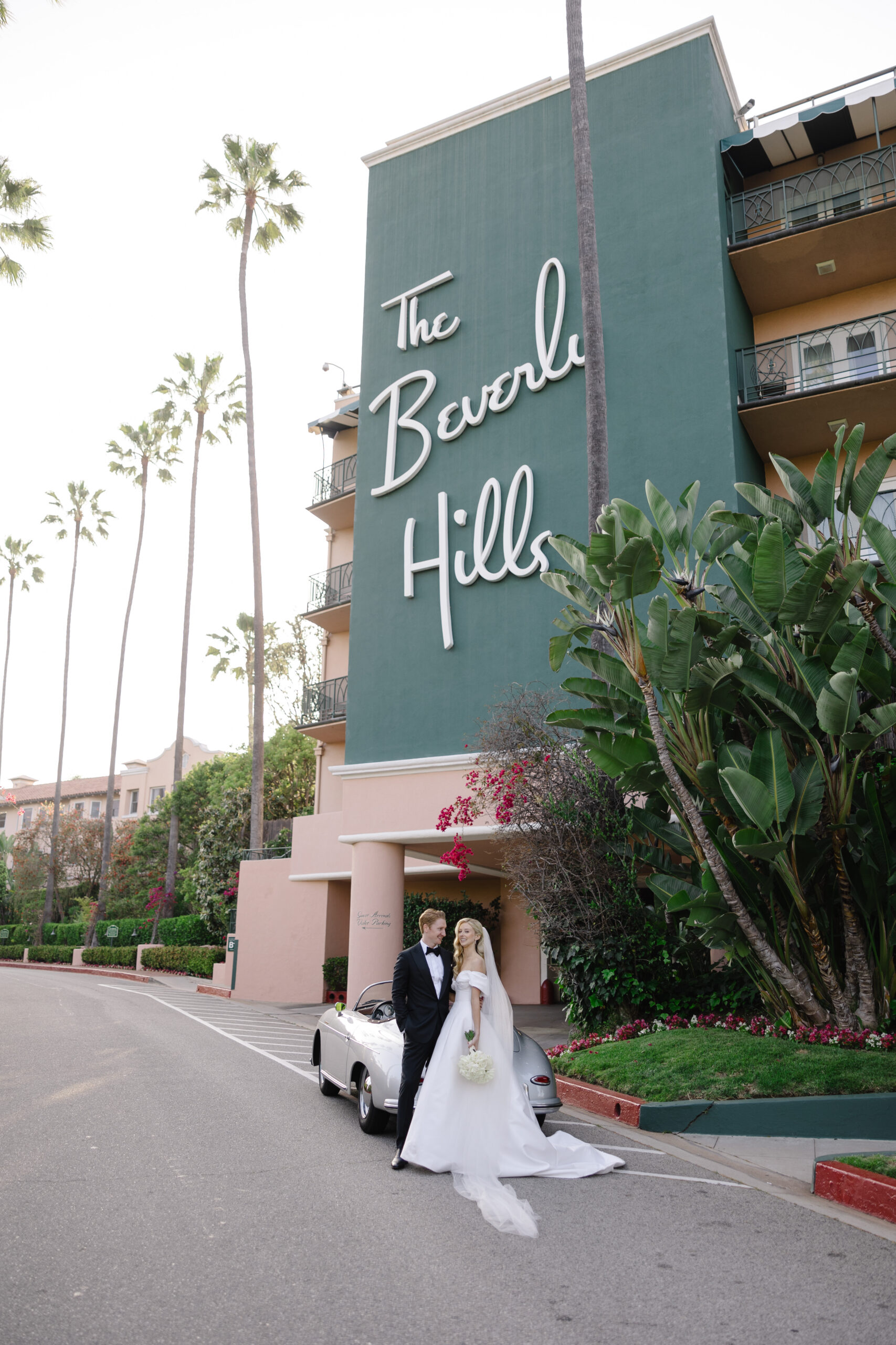 A couple in wedding attire stands in front of a silver car outside The Beverly Hills Hotel, with the hotel's large sign visible on the green exterior wall.