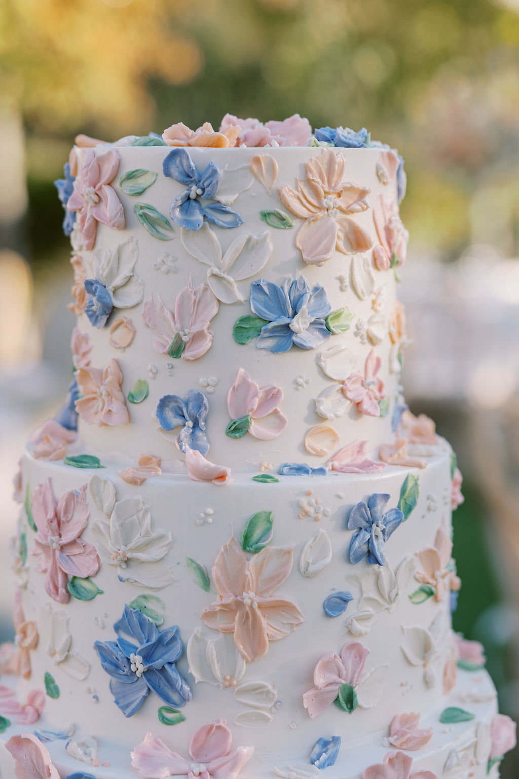A three-tiered white cake decorated with pastel pink, blue, and peach floral icing, displayed outdoors.
