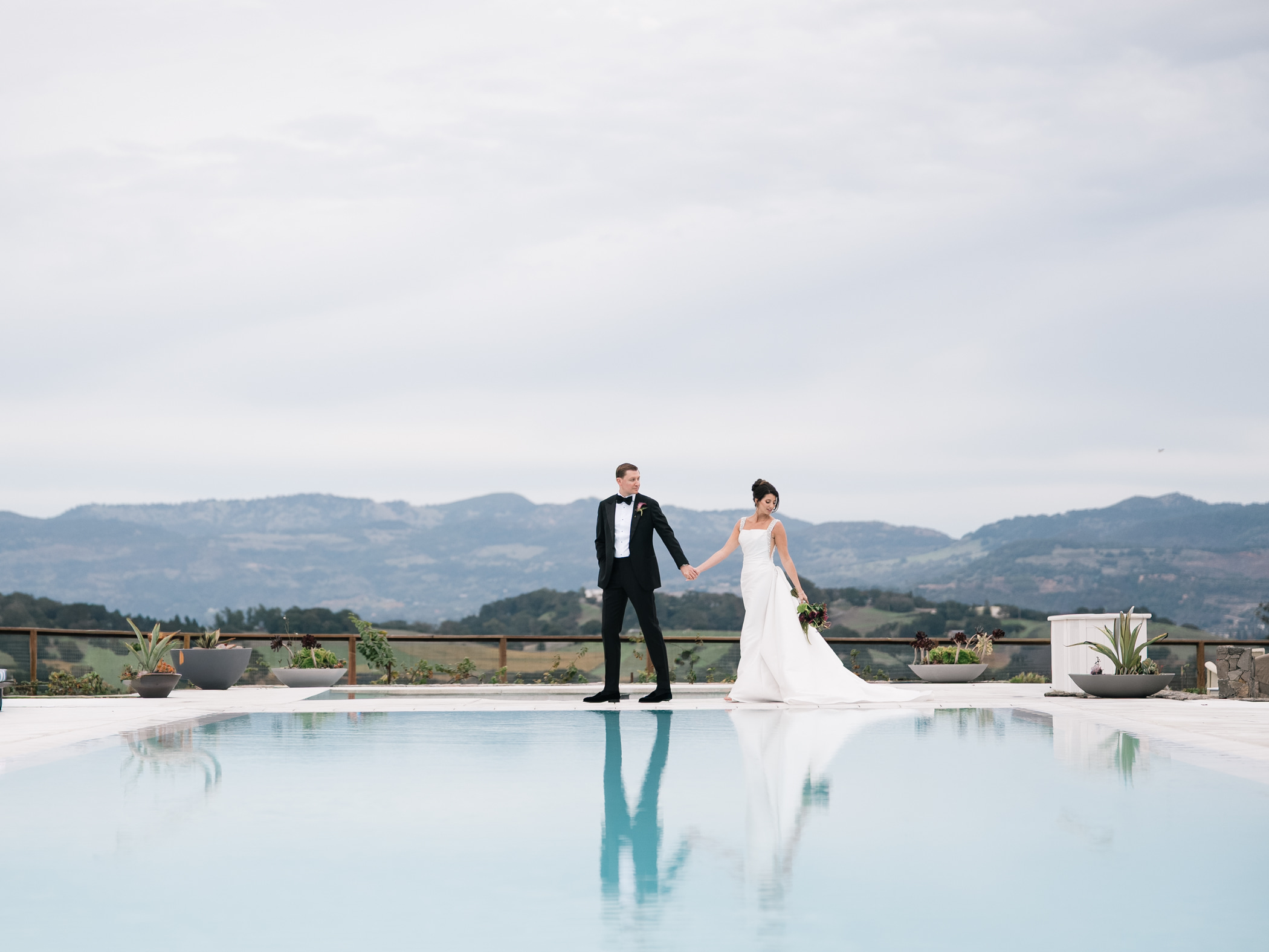 A bride and groom stand hand in hand by a pool, with mountains and cloudy sky in the background.