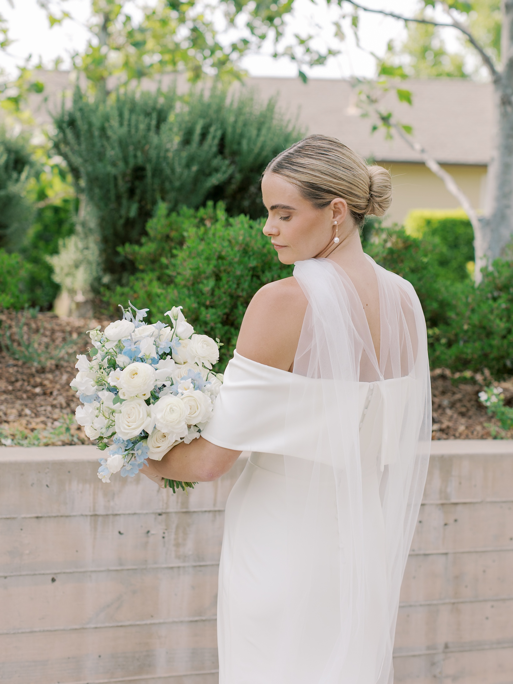 A bride in an off-the-shoulder white gown holds a bouquet of white roses and light blue flowers, standing outdoors near greenery and a low concrete wall.