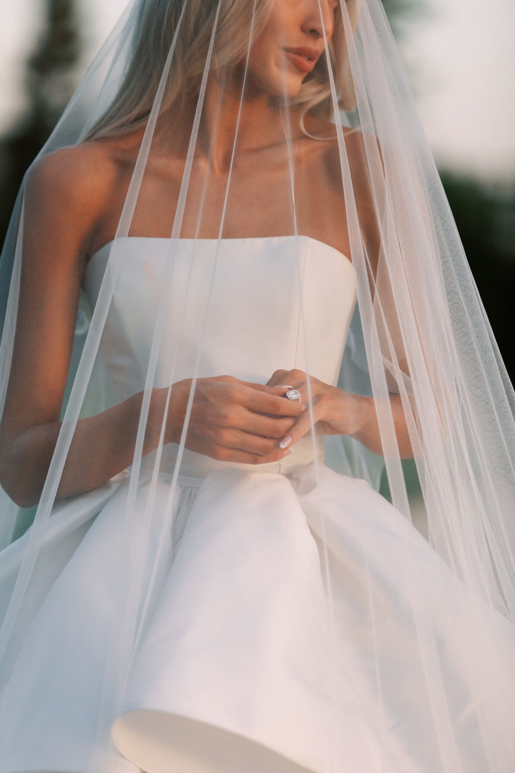 A bride in a strapless white wedding gown and veil stands outdoors, holding her hands together in front of her waist.