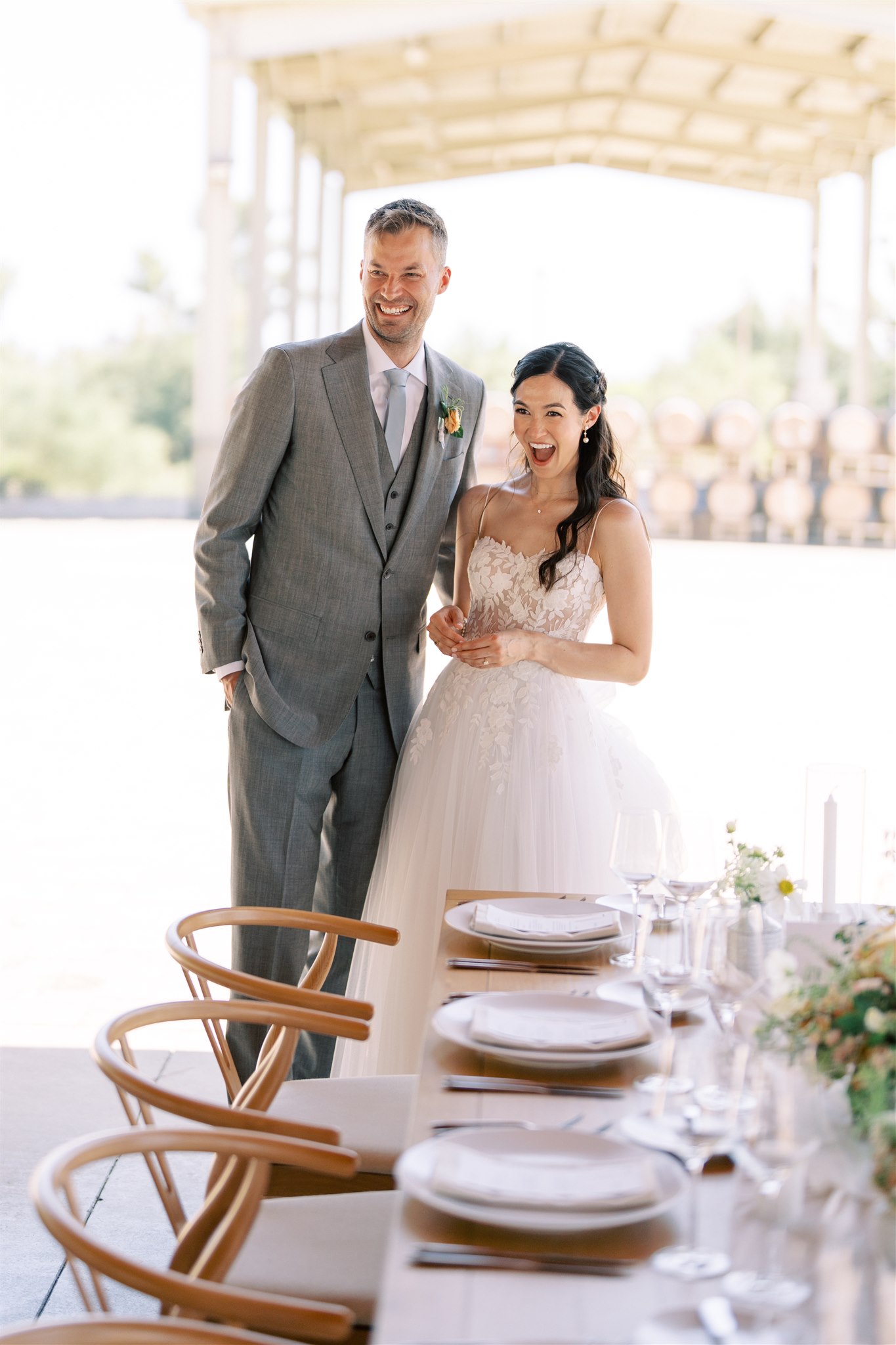 A bride and groom stand smiling and looking ahead beside a set dining table at an outdoor venue.