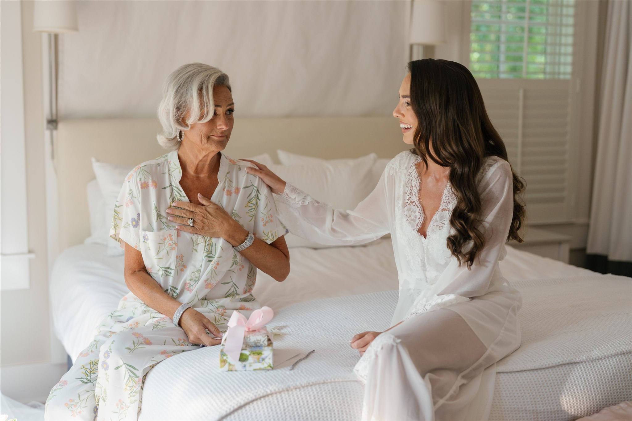 An older woman sits on a bed holding a small gift with a pink bow while a younger woman touches her shoulder; both are wearing light-colored robes.