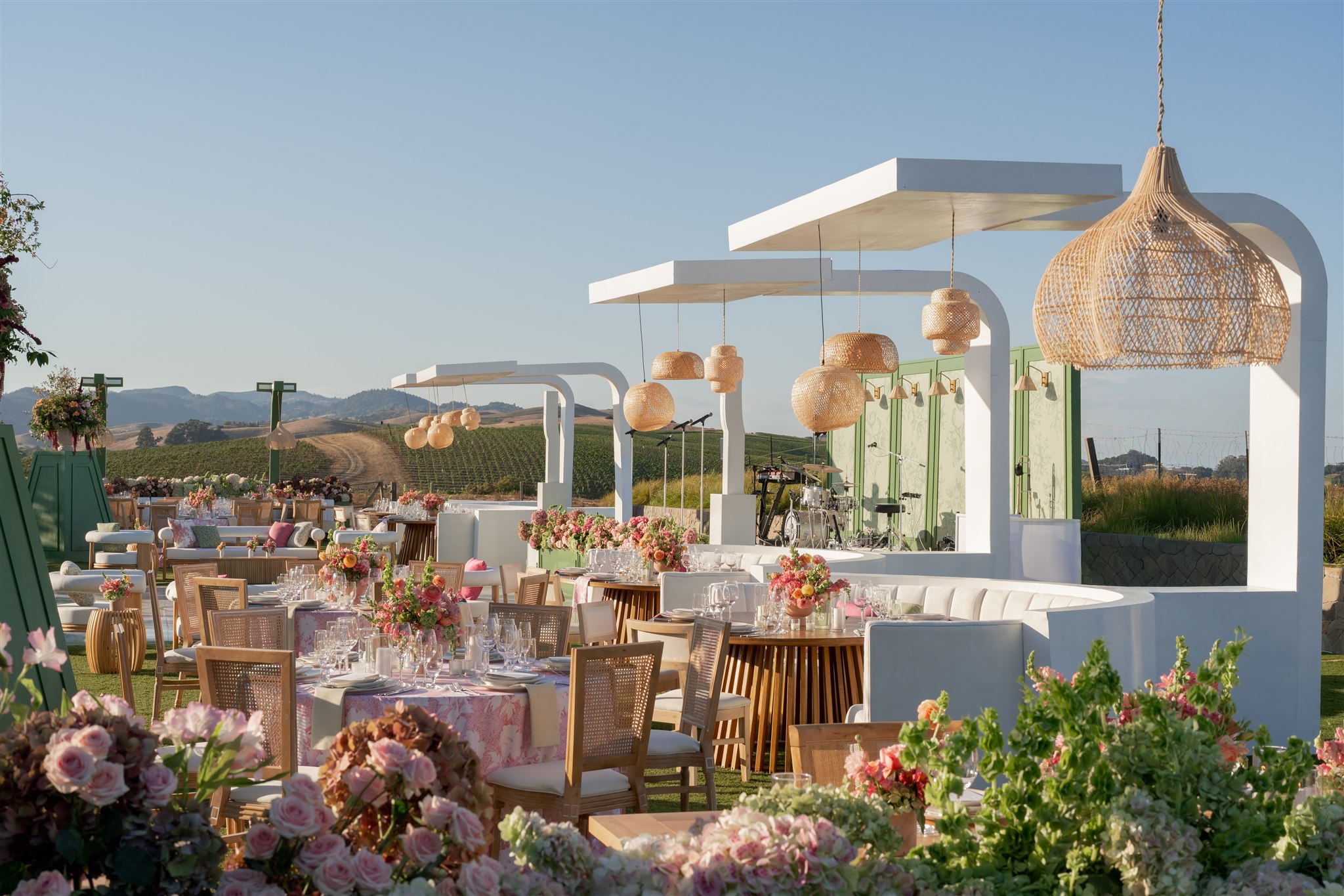 Outdoor event setup with decorated tables, floral arrangements, white canopy structures, wicker pendant lights, and a scenic vineyard in the background.