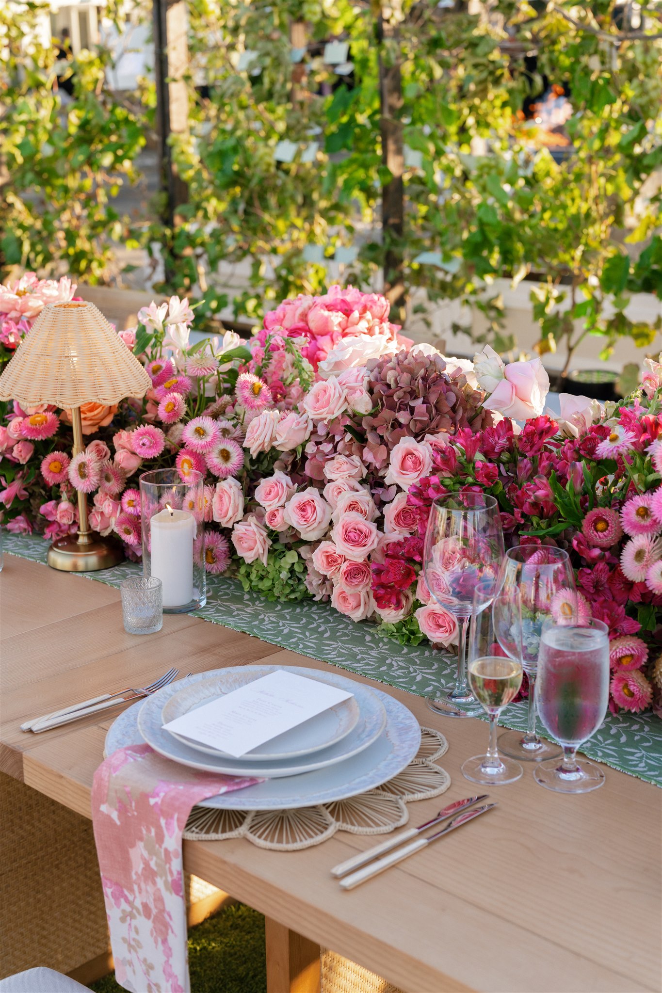 A wooden table set for dining features plates, glasses, and a menu, with a centerpiece of pink flowers and a small lamp. Greenery is visible in the background.