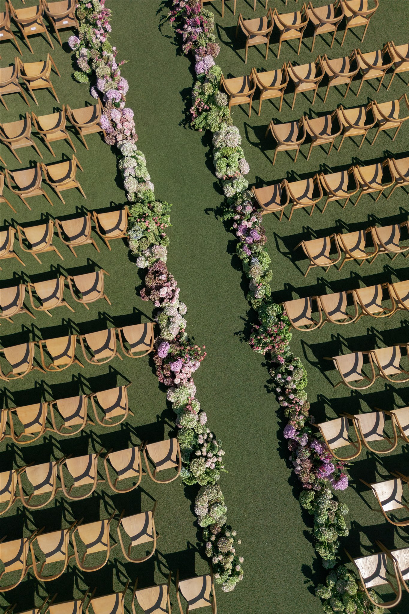 Aerial view of rows of empty wooden chairs arranged on grass with two parallel lines of assorted flowers forming an aisle in the center.
