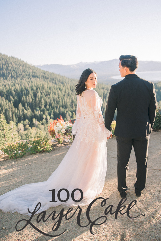5 A bride and groom stand holding hands on a scenic overlook with mountains and trees in the background, wearing formal wedding attire. "100 Layer Cake" is written at the bottom.