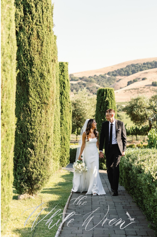 6 A bride and groom walk hand in hand along a brick path lined with tall green hedges, with hills and trees in the background.